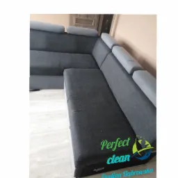 Szary narożnik z wysuwaną skrzynią na pościel, na podłodze panele, widoczne logo firmy sprzątającej 'Perfect clean Ewelina Dąbrowska'.