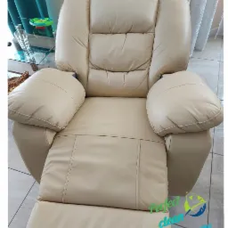 Beżowy, skórzany fotel relaksacyjny z wysuniętym podnóżkiem, widok z przodu, na podłodze z płytek. W tle stolik z blatem ze szkła i zasłony.