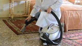 Pracownik w białym uniformie odkurza wzorzysty dywan na podłodze z terakoty za pomocą profesjonalnego odkurzacza piorącego z metalową obudową, widoczny fragment łóżka z beżową narzutą.