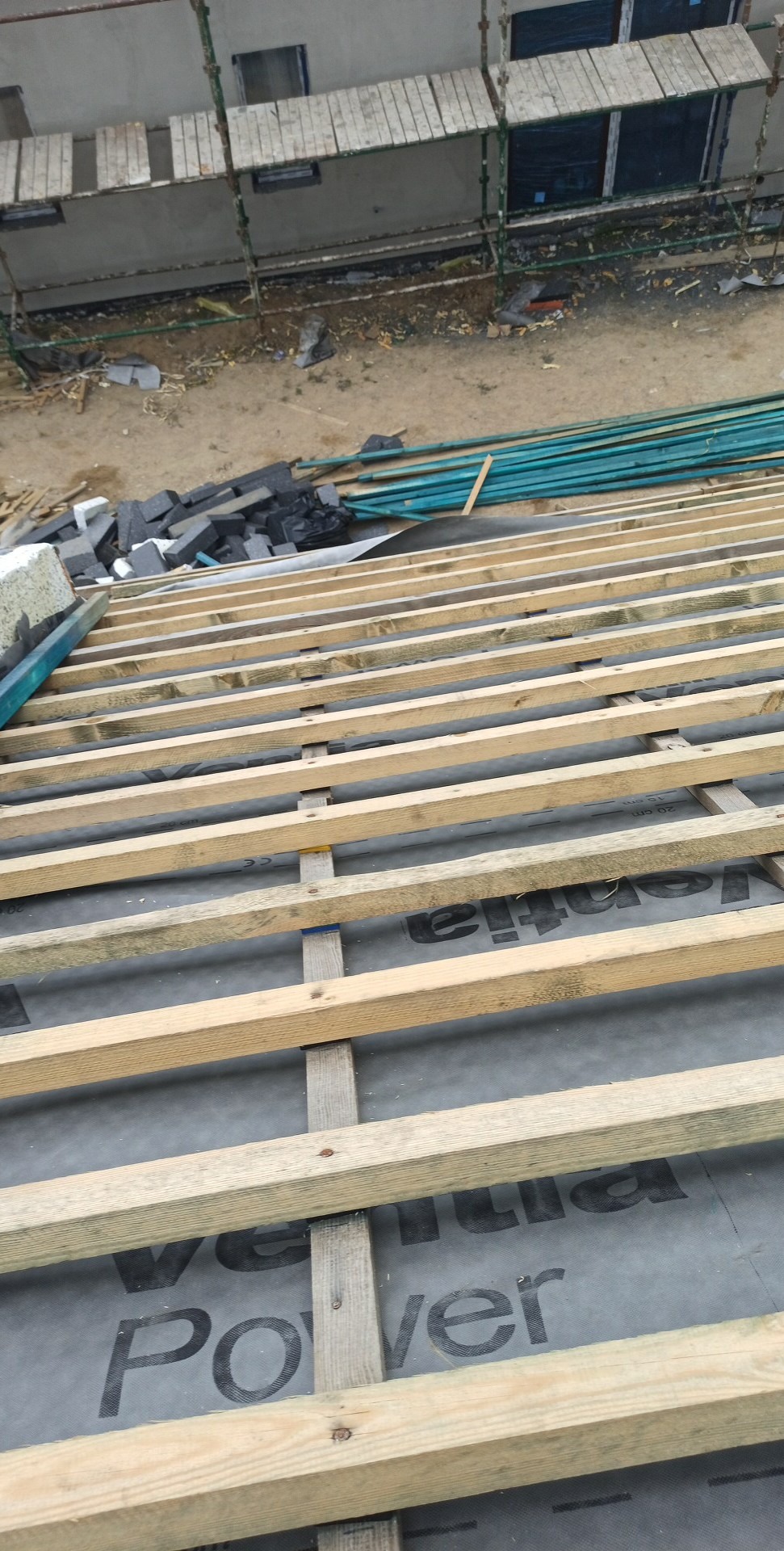 Konstrukcja drewniana dachu z widoczną membraną dachową i przygotowanymi płytkami dachowymi w tle, rusztowanie przy budynku.