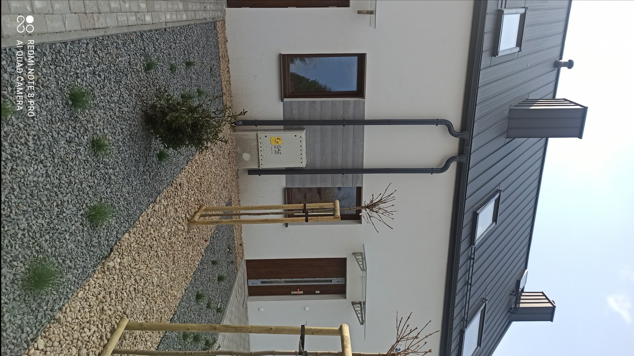 Nowoczesny dom jednorodzinny z ciemnoszarym dachem z blachy na rąbek stojący, widoczne okna dachowe i rynny w kolorze dachu, elewacja biała z elementami dekoracyjnymi imitującymi drewno, obok...