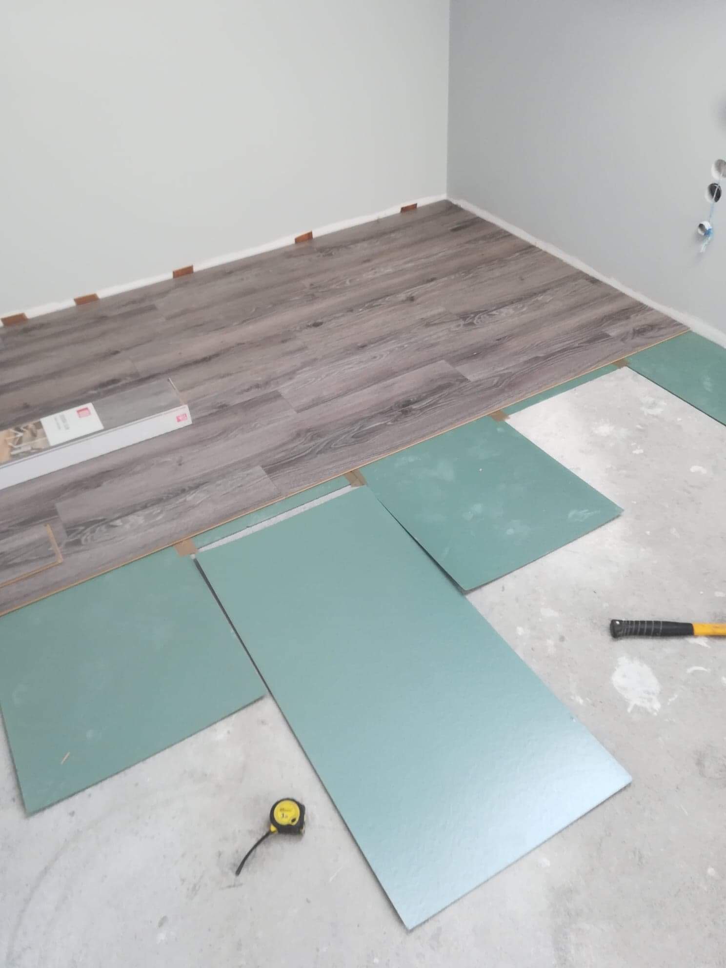 Proces montażu paneli podłogowych: częściowo ułożona podłoga w odcieniach szarości, zielone płyty podkładowe, miarka i młotek w pomieszczeniu o jasnych ścianach.
