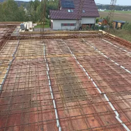 Zbrojenie stropu z widocznymi prętami stalowymi tworzącymi siatkę, szalunek z desek i dystanse betonowe, w tle dom z panelami słonecznymi.