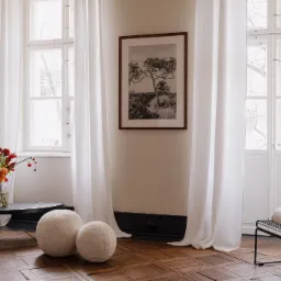 Jasne, zwiewne zasłony z lnu zdobią okna w pokoju z parkietem, uzupełnione czarno-białą fotografią drzewa w brązowej ramie, bukietem kwiatów w wazonie na niskim stoliku i dekoracyjnymi kulami...