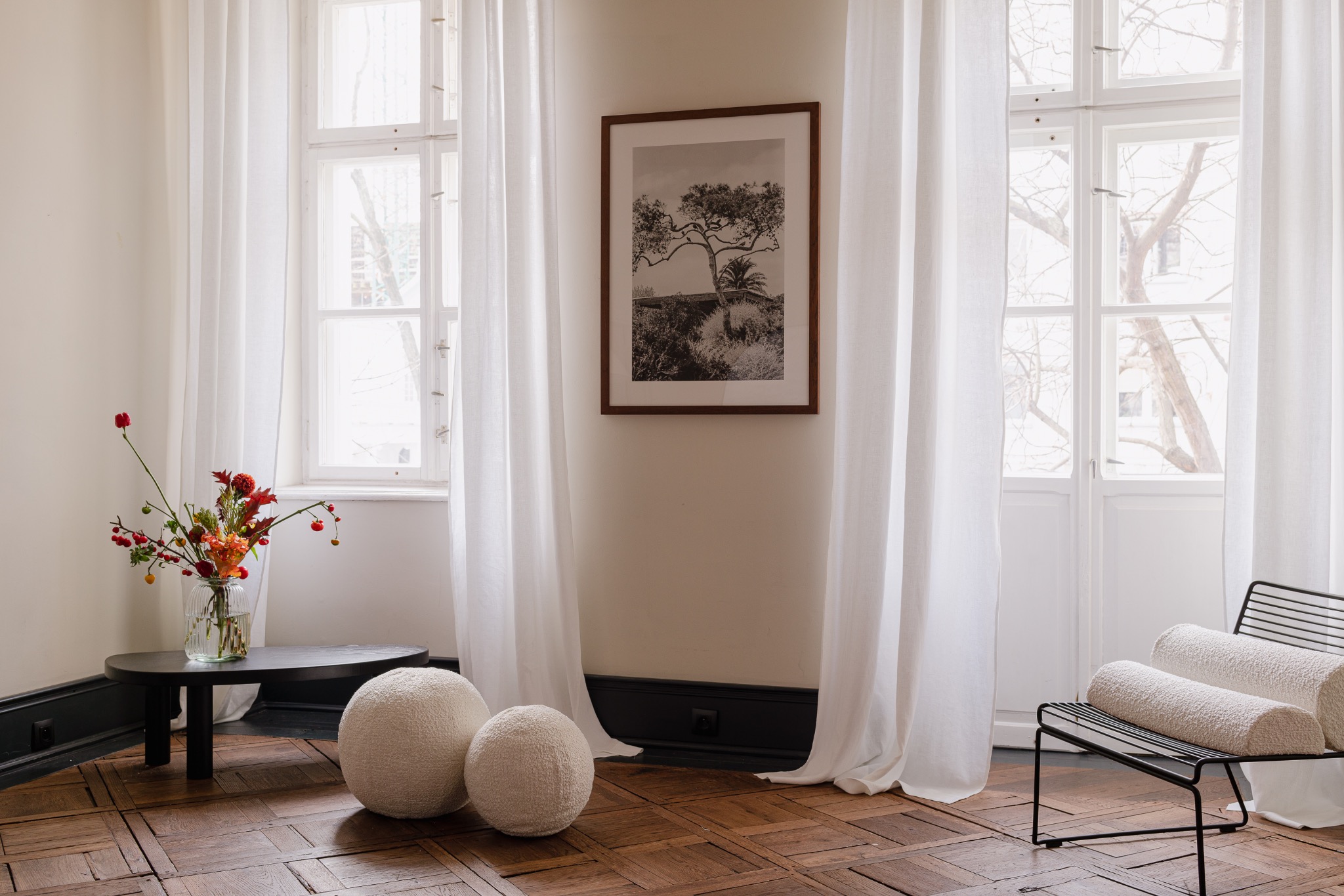Jasne, zwiewne zasłony z lnu zdobią okna w pokoju z parkietem, uzupełnione czarno-białą fotografią drzewa w brązowej ramie, bukietem kwiatów w wazonie na niskim stoliku i dekoracyjnymi kulami...