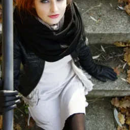 Portret kobiety z rudymi włosami w czarnej skórzanej kurtce, szaliku i rękawiczkach, siedzącej na betonowych schodach z opadłymi liśćmi.