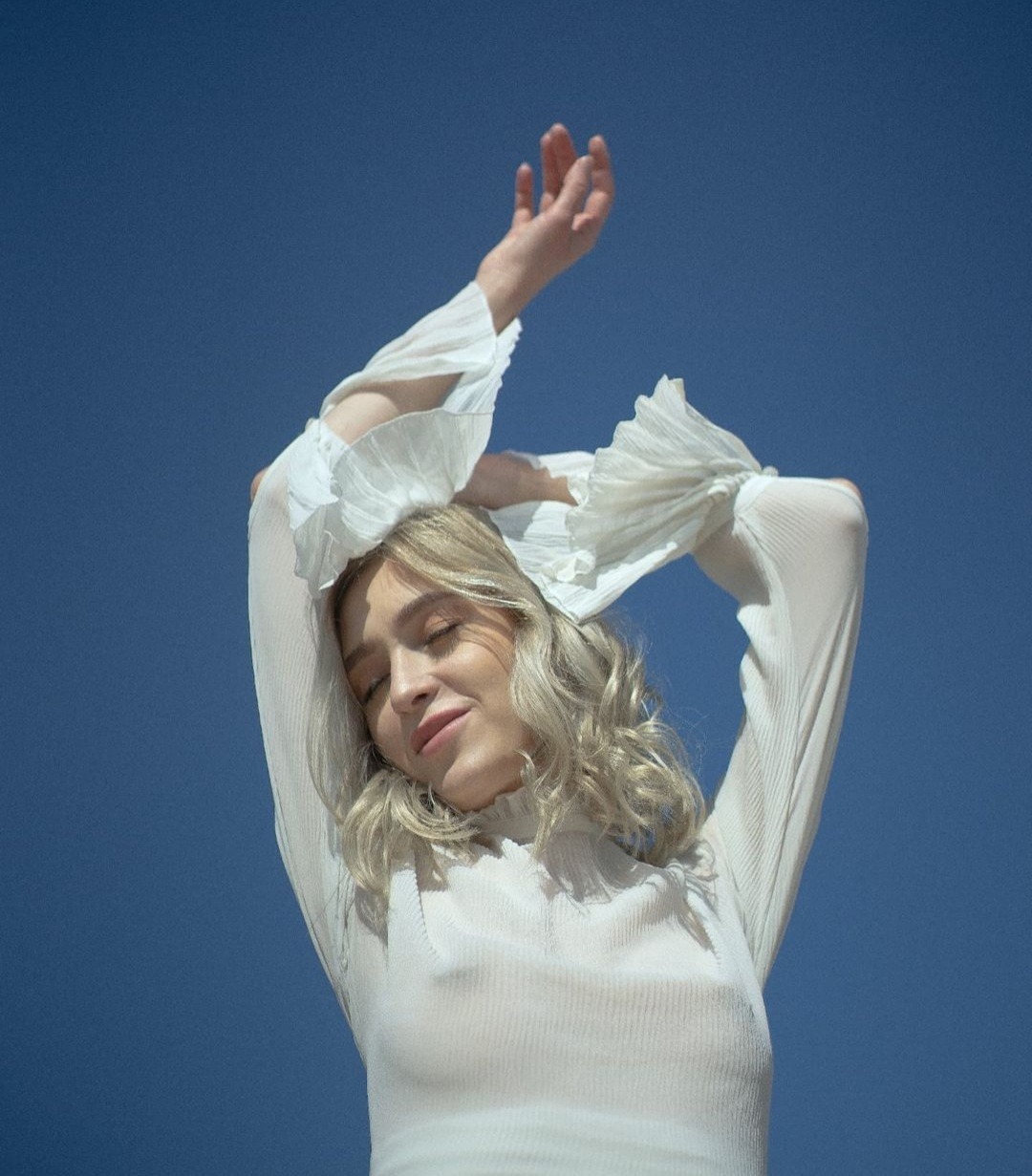 Portret kobiety z blond włosami, ubrana w białą, prześwitującą bluzkę z falbanami na rękawach, pozująca na tle błękitnego nieba.
