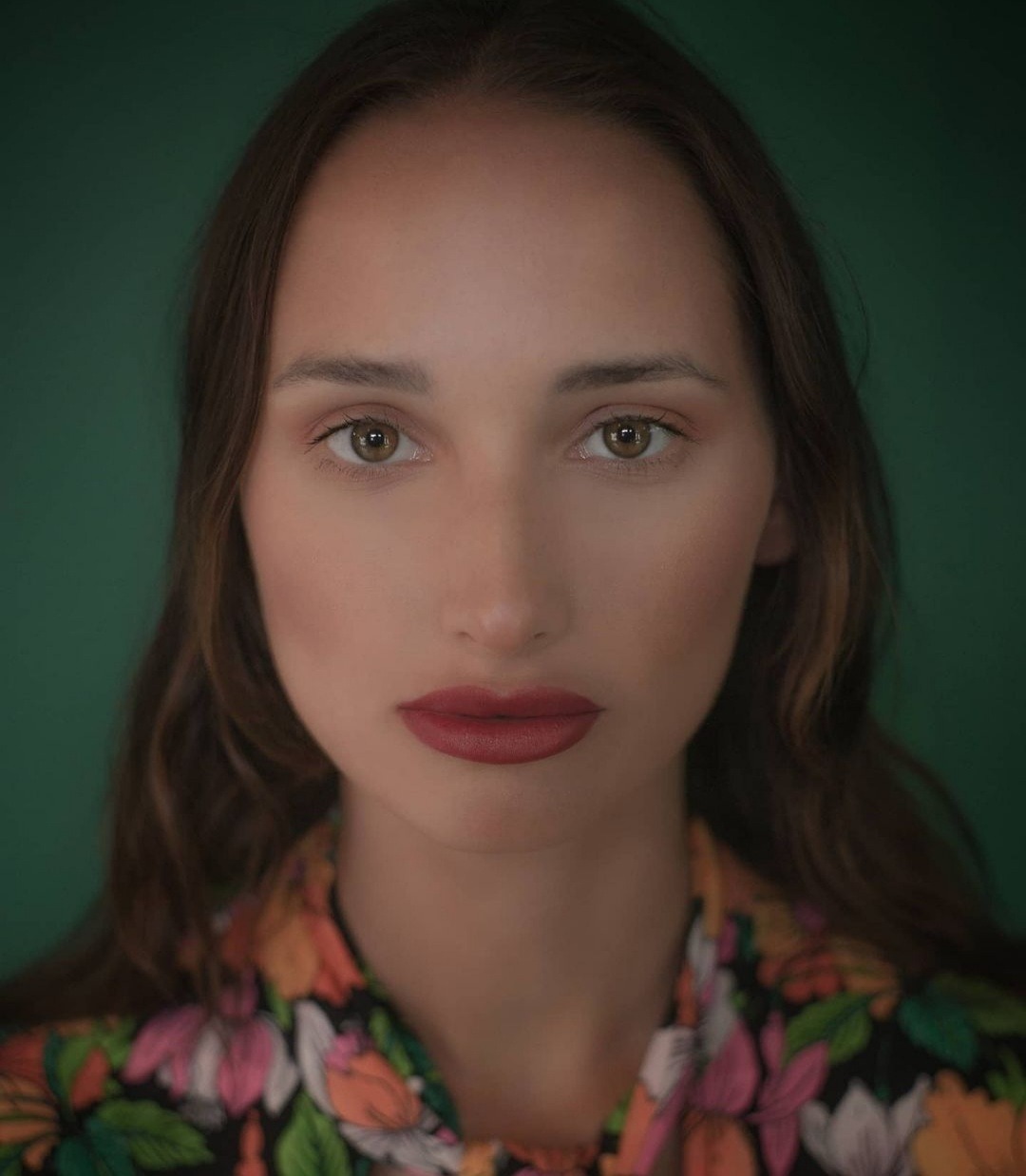 Portret kobiety z długimi brązowymi włosami, zielonymi oczami i ciemnoczerwoną szminką, ubrana w bluzkę w kwiaty na tle ciemnozielonego tła.