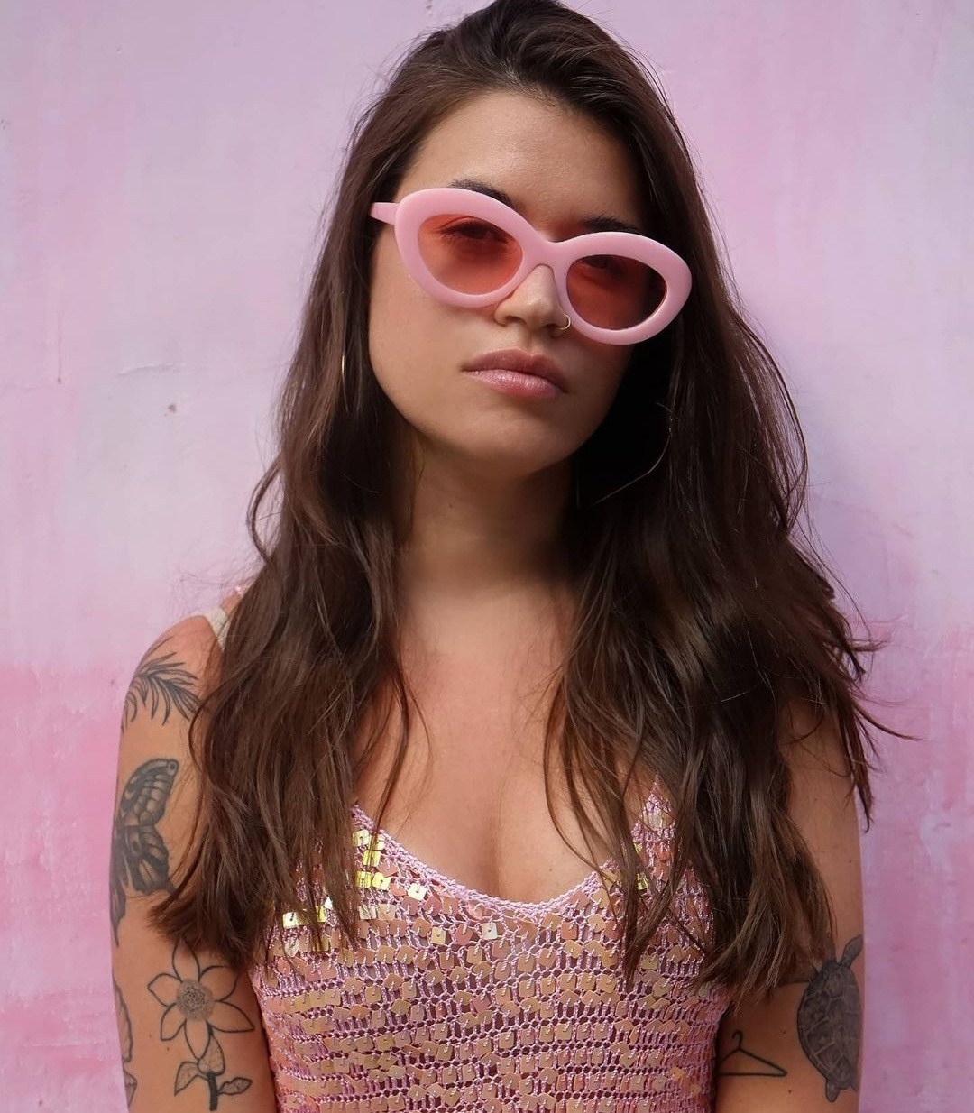 Portret młodej kobiety w różowych okularach i błyszczącej, ażurowej bluzce na tle różowej ściany, z widocznymi tatuażami na ramionach.