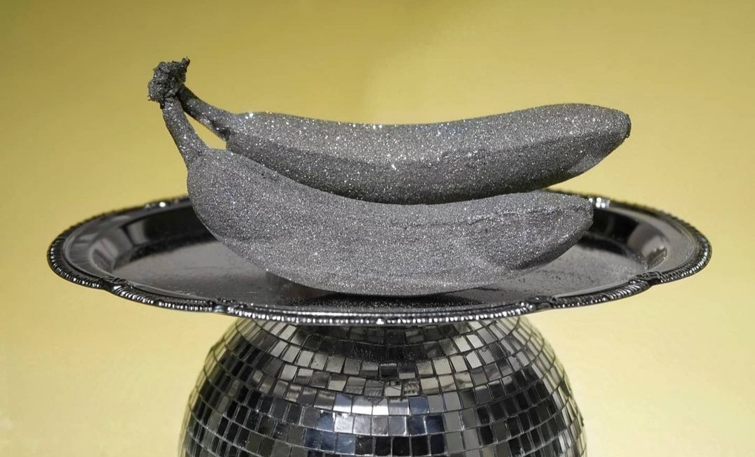 Dwie banany pokryte srebrnym brokatem leżące na czarnym talerzu, całość umieszczona na podstawie z lustrzanych kwadratów.