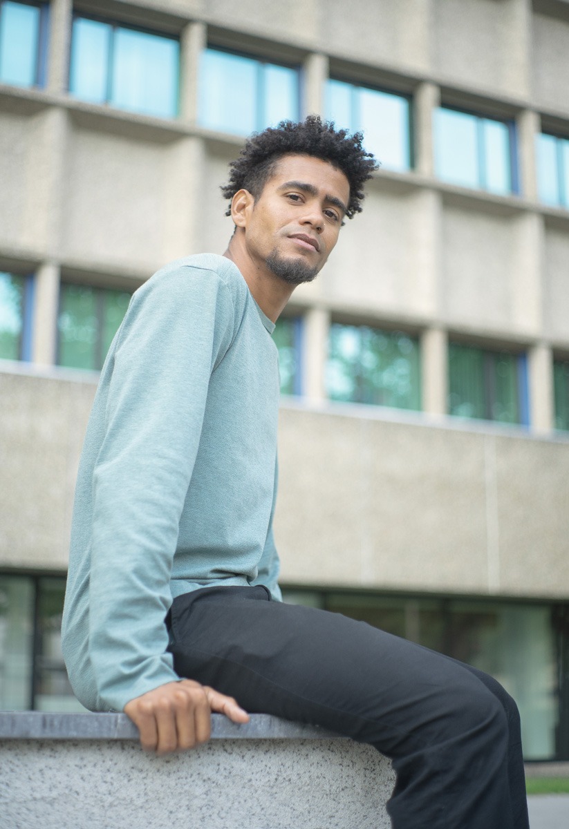 Młody mężczyzna w jasnej bluzie i czarnych spodniach siedzi na murku na tle modernistycznego budynku z dużymi oknami.