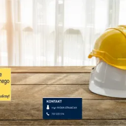 Dwa kaski ochronne, żółty i biały, ułożone jeden na drugim na drewnianym stole, w tle ulotka z tekstem 'Doradztwo w zakresie organizacji bezpiecznego miejsca pracy' i dane kontaktowe.