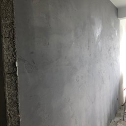 DOMBUD DOMINIK WLAZŁO - Ściana wewnętrzna pokryta szarą, dekoracyjną fakturą, widoczne nierówności i wzory na powierzchni, surowy betonowy narożnik, fragment podłogi w trakcie remontu.