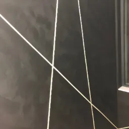 Ciemna ściana z dekoracyjnym efektem i geometrycznymi, jasnymi liniami, widoczny fragment okna i zabezpieczenie podłogi.