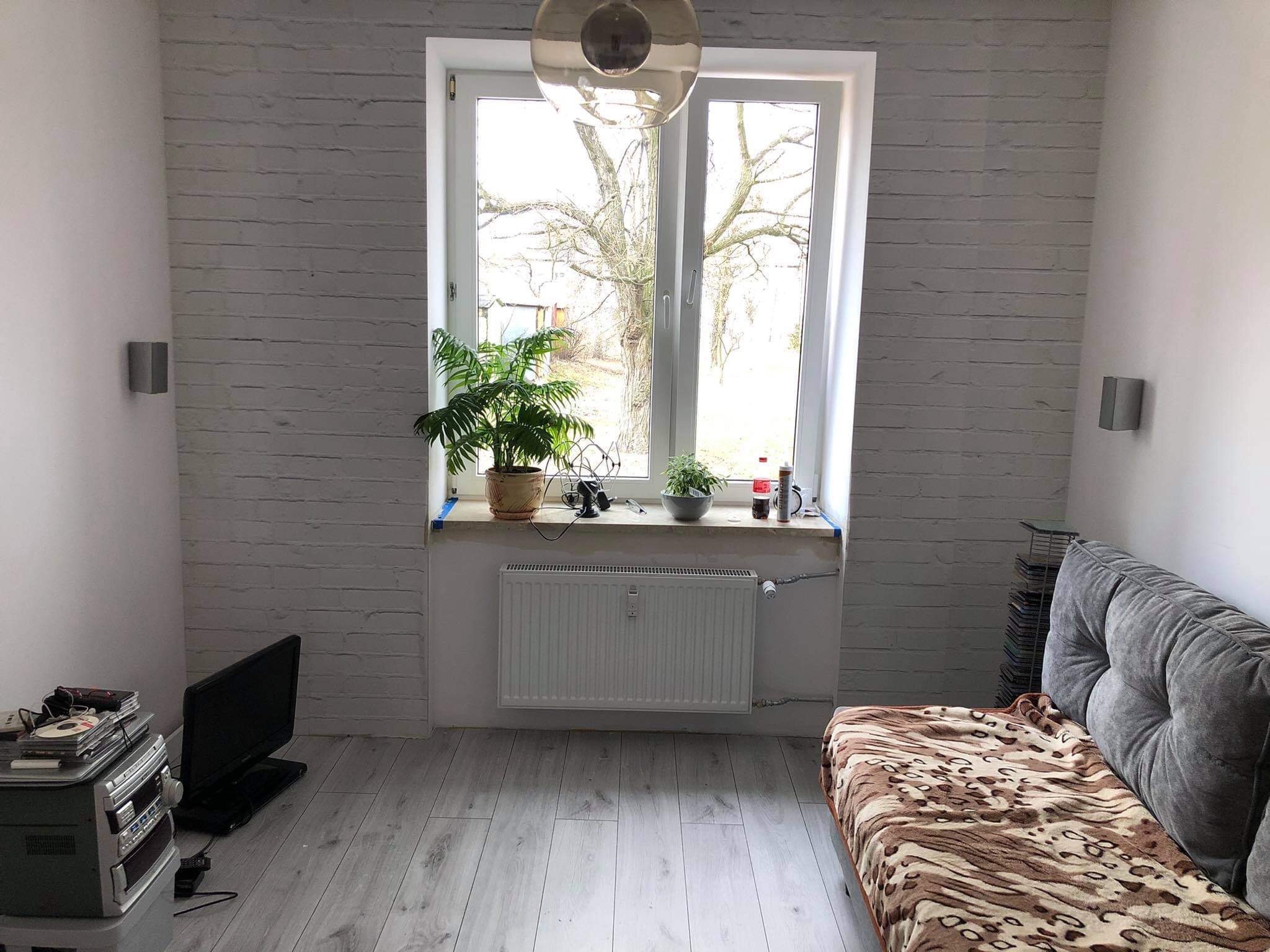 Pokój po odświeżeniu: ściana z białej cegły, okno z widokiem na drzewo, szara sofa z narzutą w panterkę, szare panele na podłodze, grzejnik pod oknem, lampa sufitowa w kształcie kuli.
