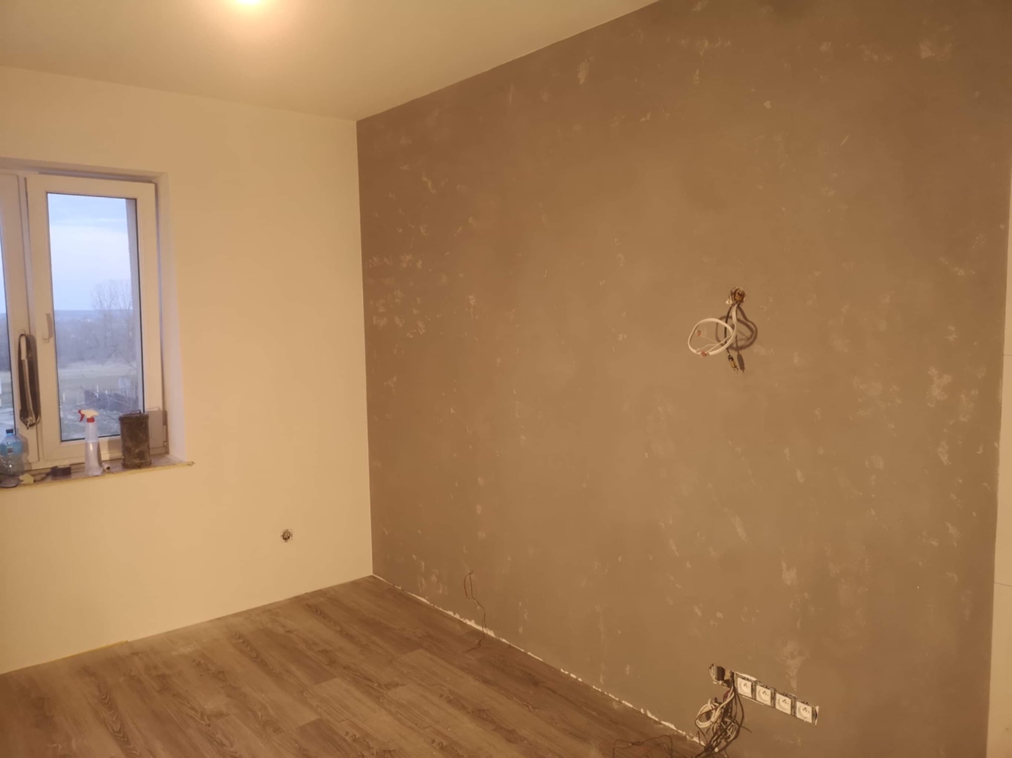 Pokój w trakcie remontu: ściana z nałożoną gładzią w odcieniach beżu, widoczne kable elektryczne, podłoga wyłożona panelami drewnopodobnymi, okno z widokiem na zewnątrz.