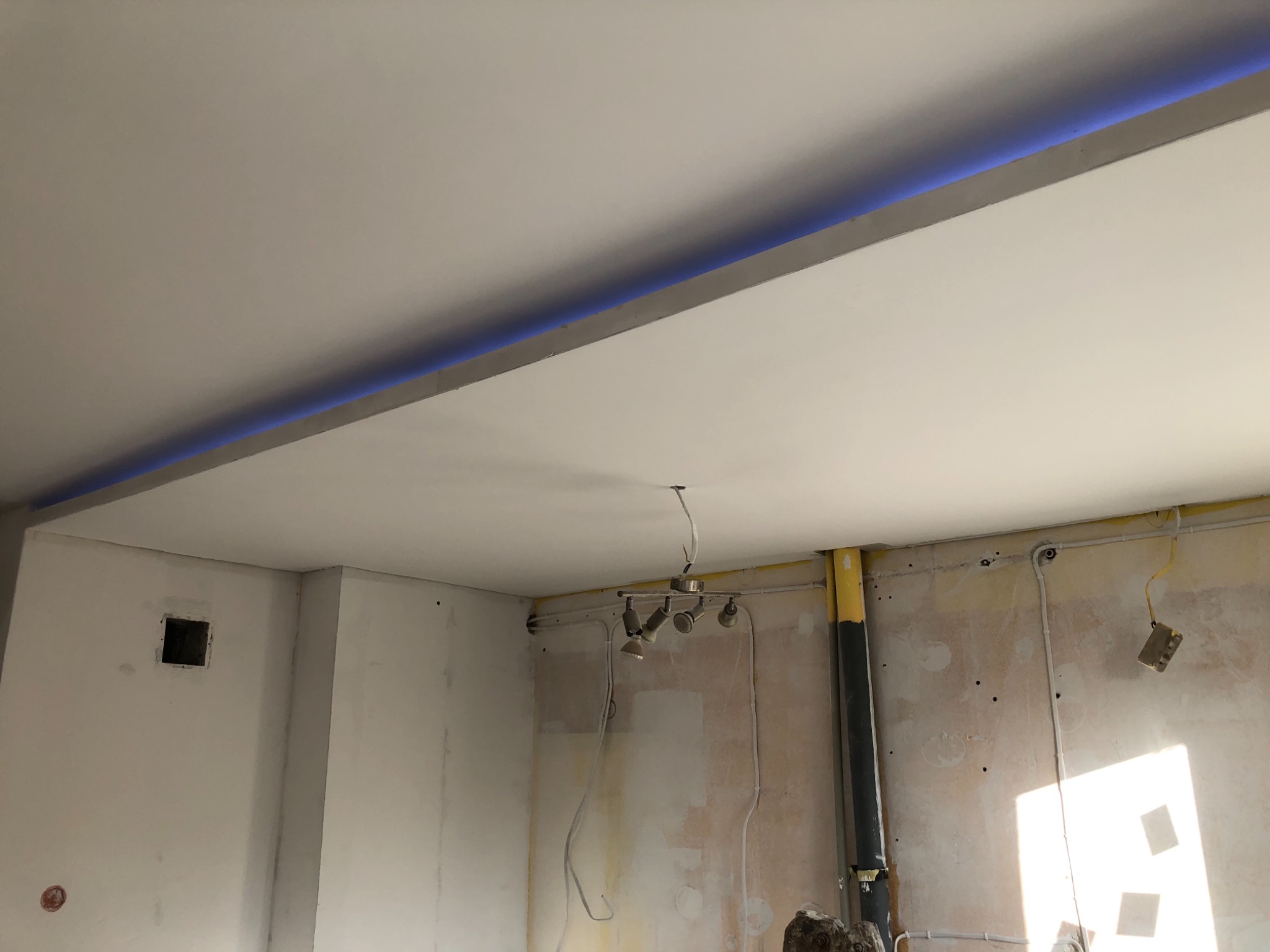 Wnętrze w trakcie remontu: gładzie na ścianach i suficie, widoczne kable i rury, sufit z oświetleniem LED w górnej części.