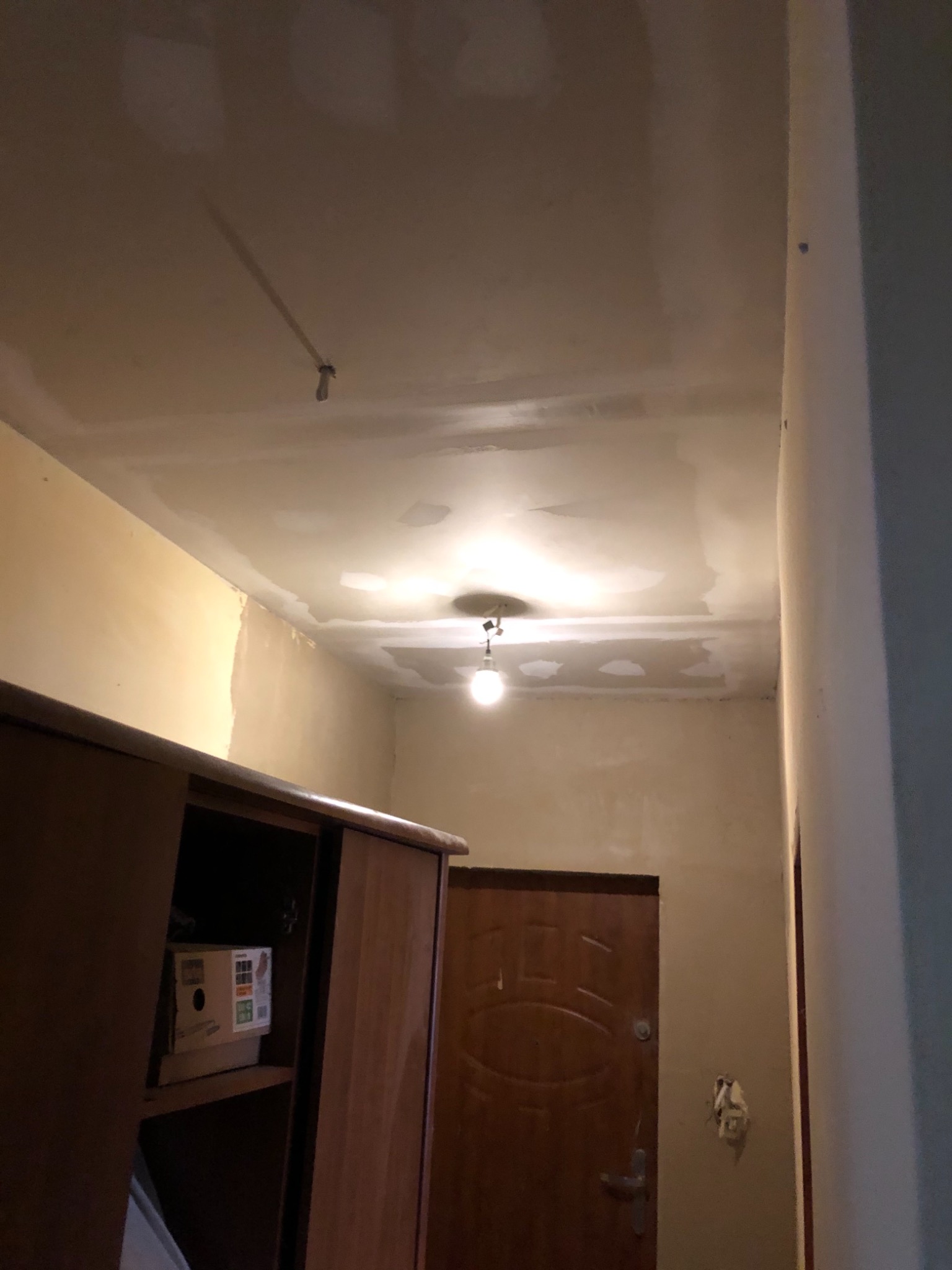 Wnętrze w trakcie prac wykończeniowych: sufit z nałożonymi gładziami, ściany częściowo pomalowane, widoczna wisząca żarówka i szafa z przesuwnymi drzwiami.