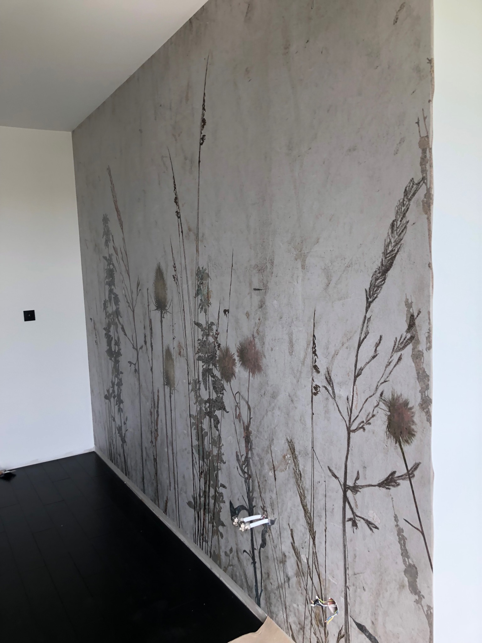 Ściana z tapetą w motywy roślinne, imitująca starą, lekko zniszczoną powierzchnię, z widocznymi przewodami elektrycznymi wystającymi ze ściany, oraz fragment czarnej podłogi.