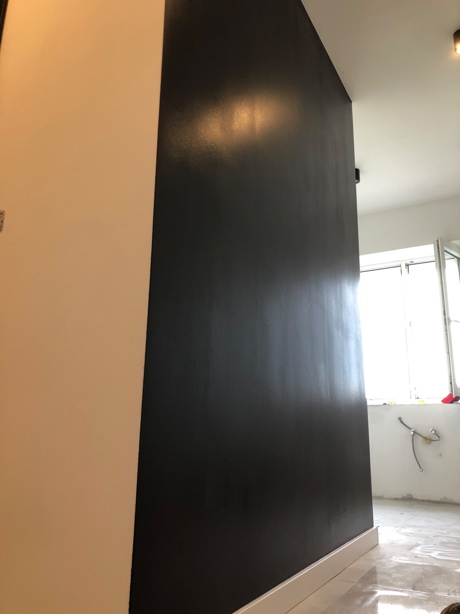 Świeżo pomalowana, błyszcząca, ciemna ściana w pomieszczeniu w trakcie remontu, widoczne białe listwy przypodłogowe, fragment ściany w kolorze kremowym, okno i instalacja wodna w tle.