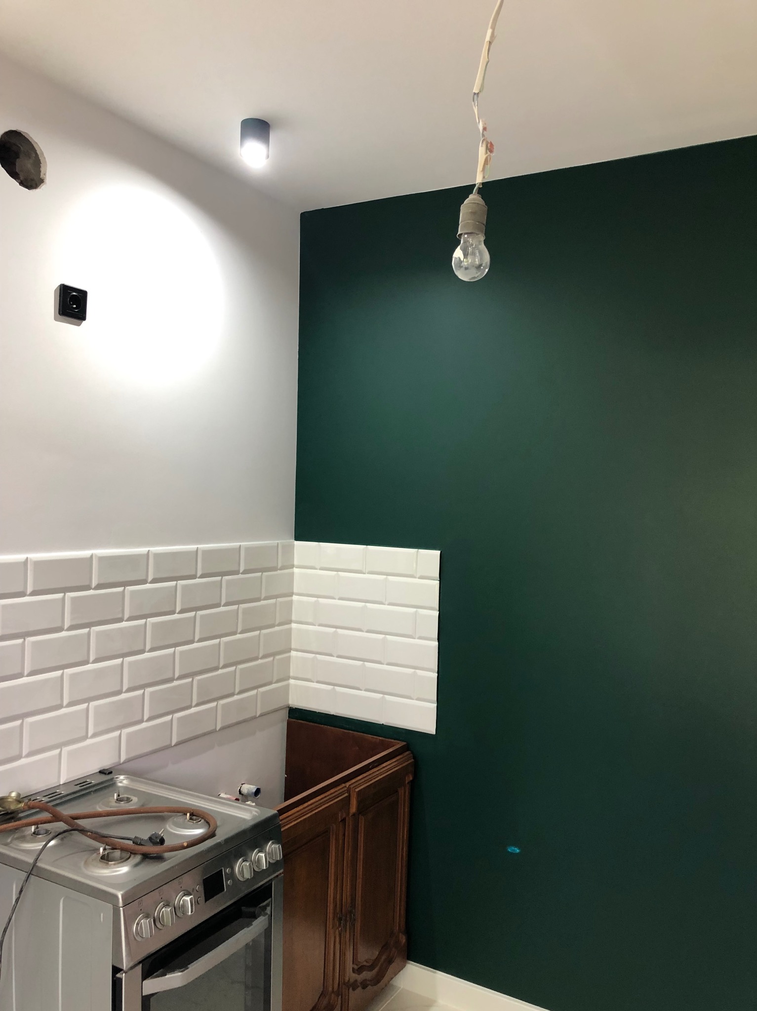Kuchnia w trakcie remontu: ściana pomalowana na butelkową zieleń, białe kafelki metra na ścianie, surowa żarówka zwisająca z sufitu, kuchenka gazowa na pierwszym planie, brak frontu szafki pod zlewem.