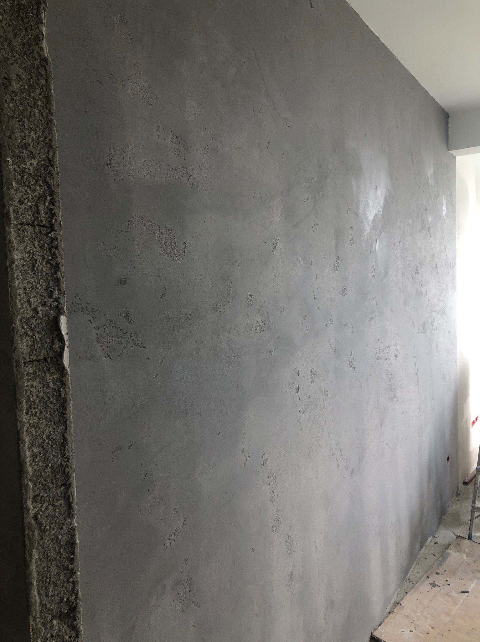 Ściana wewnętrzna pokryta szarą, dekoracyjną fakturą, widoczne nierówności i wzory na powierzchni, surowy betonowy narożnik, fragment podłogi w trakcie remontu.