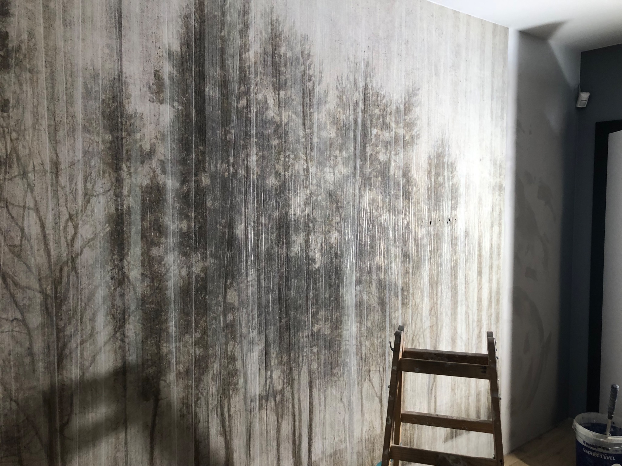 Tapeta z motywem lasu na ścianie, drabina malarska w prawym dolnym rogu, wiadro z farbą i pędzlem widoczne obok, surowy, artystyczny efekt.