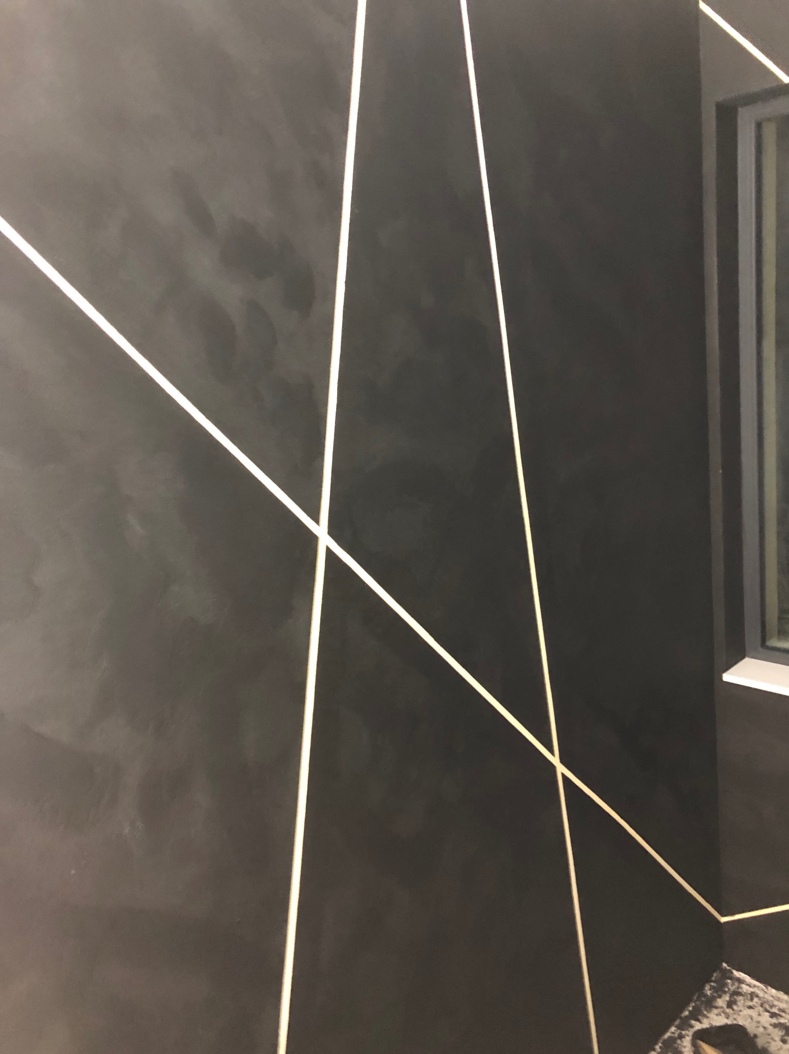 Ciemna ściana z dekoracyjnym efektem i geometrycznymi, jasnymi liniami, widoczny fragment okna i zabezpieczenie podłogi.