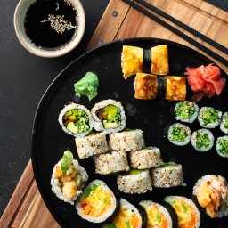 Sushi Kado - Zestaw różnorodnych sushi na czarnym talerzu, obok miseczka z sosem sojowym posypanym sezamem, pałeczki i drewniana deska do krojenia.