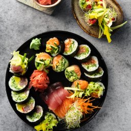 Sushi Kado - Zestaw sushi i sashimi z dodatkami, ułożony na czarnym talerzu, obok miseczka z marynowanym imbirem i rolki w papierze ryżowym na brązowym talerzu, całość na szarym tle.