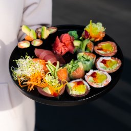 Sushi Kado - Osoba trzyma czarny talerz z różnorodnym zestawem sushi i sashimi, w tym rolki z łososiem, tuńczykiem, awokado i warzywami, udekorowane imbirem i kiełkami.