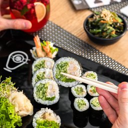 Sushi Kado - Czarny talerz z sushi maki z krewetką i ogórkiem, pałeczki w dłoni, napój owocowy z miętą i limonką oraz sałatka z wodorostów w tle.