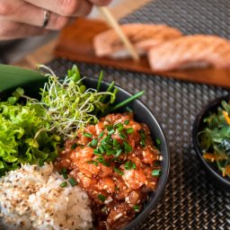 Sushi Kado - Poke bowl z łososiem, ryżem, sałatą, kiełkami i szczypiorkiem w czarnej misce, trzymana pałeczkami przez osobę z obrączką, na tle deski z łososiem i miski z wodorostami.