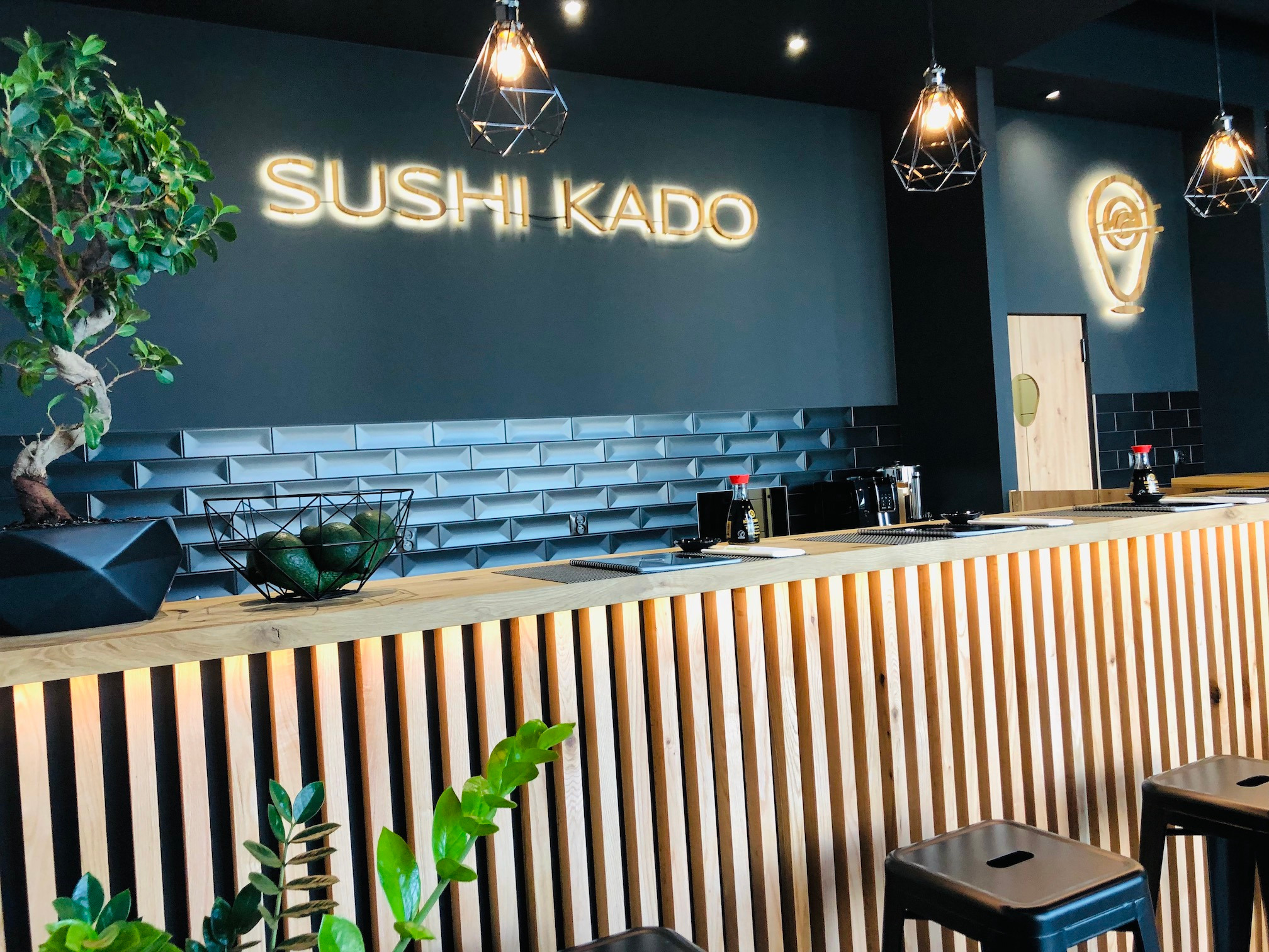 Wnętrze restauracji sushi z neonowym napisem 'SUSHI KADO' na szarej ścianie, drewnianym barem, dekoracyjnymi lampami i roślinami w doniczkach.