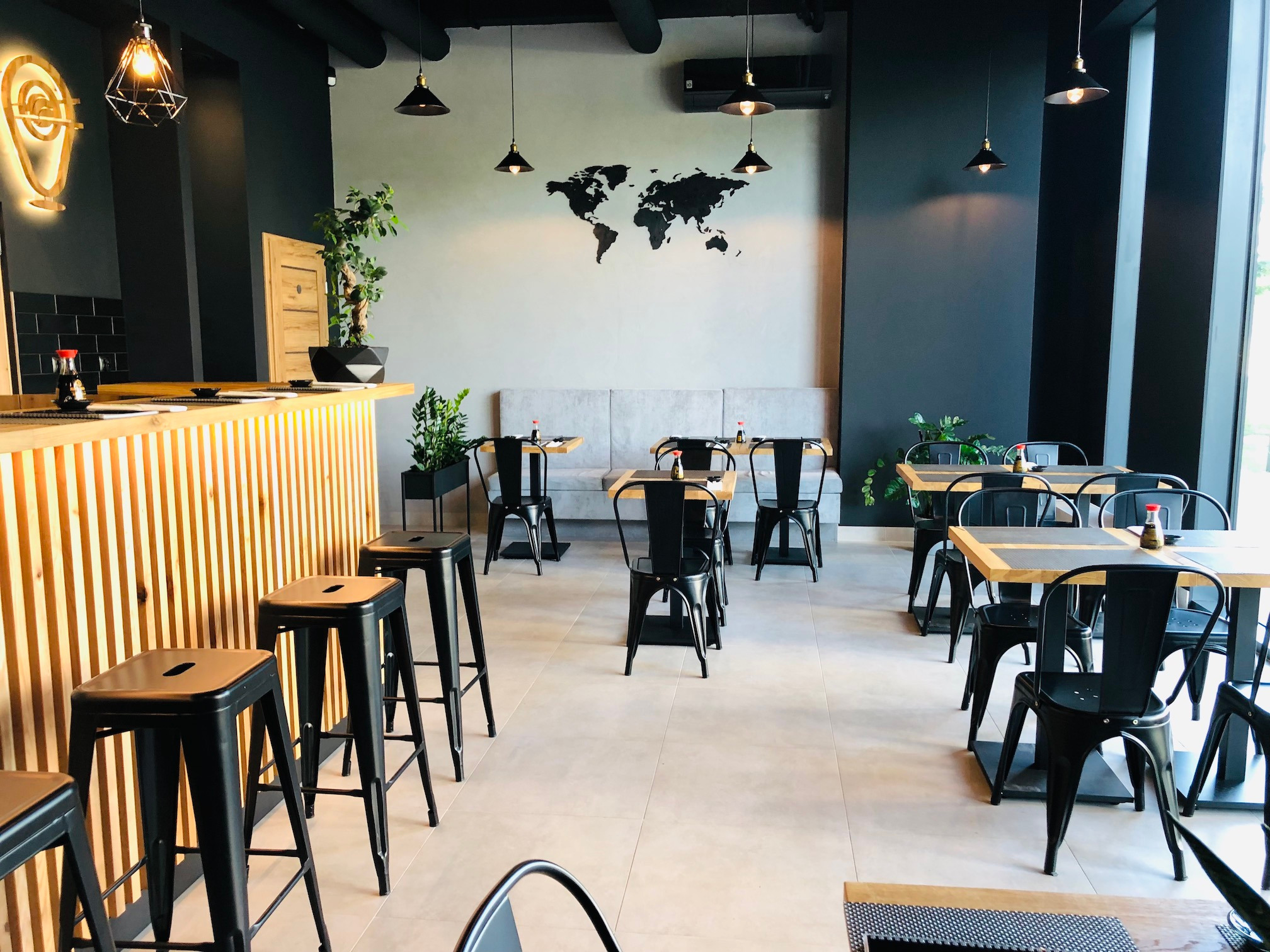 Wnętrze nowoczesnej restauracji z drewnianym barem, czarnymi krzesłami i stołami, dekoracją w postaci mapy świata na ścianie i wiszącymi lampami.