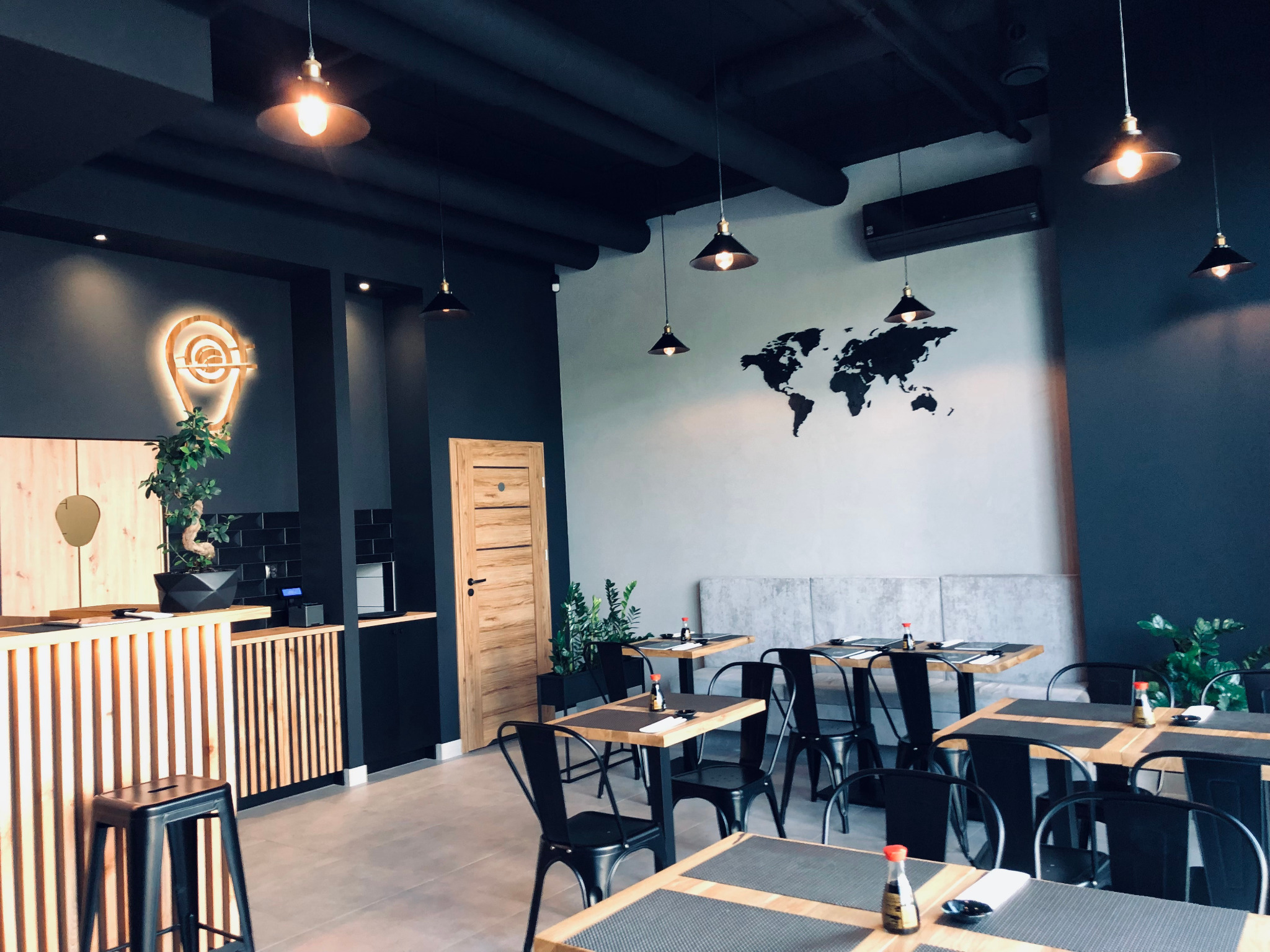 Wnętrze restauracji w stylu minimalistycznym z drewnianymi stolikami nakrytymi dla gości, dekoracyjna mapa świata na ścianie i podświetlane logo firmy w kształcie pinezki.