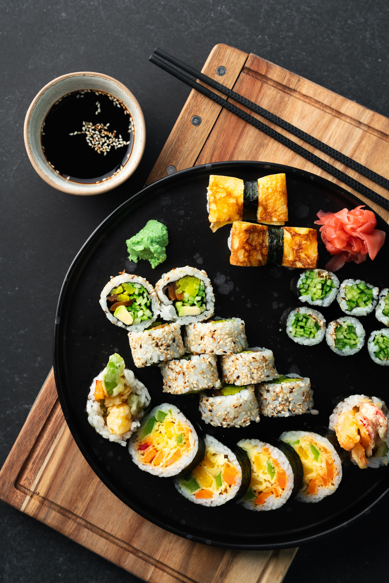 Zestaw różnorodnych sushi na czarnym talerzu, obok miseczka z sosem sojowym posypanym sezamem, pałeczki i drewniana deska do krojenia.