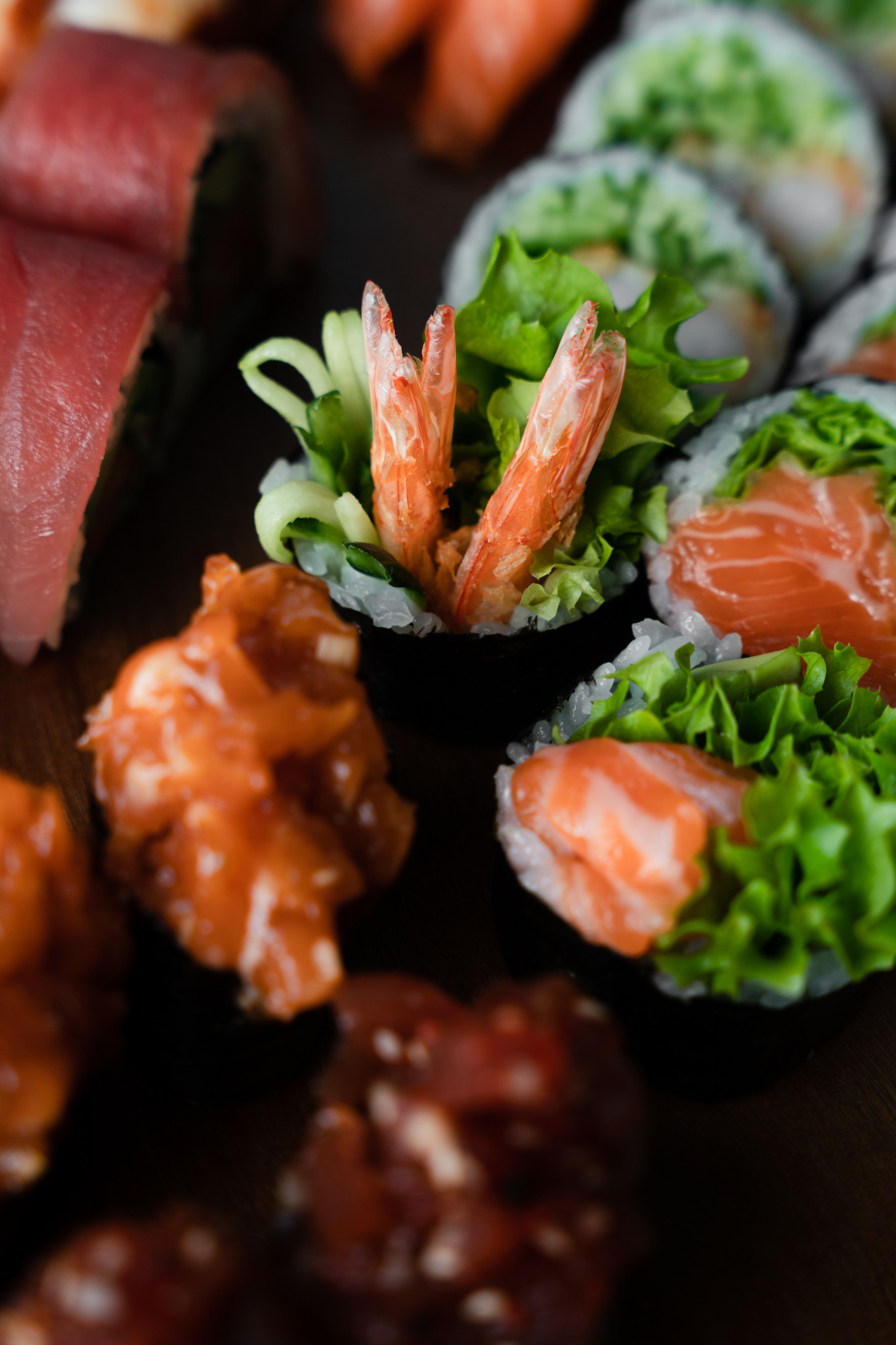 Różnorodne sushi: rolki z tuńczykiem, łososiem i krewetkami, udekorowane sałatą i ogórkiem, podane na ciemnym tle.