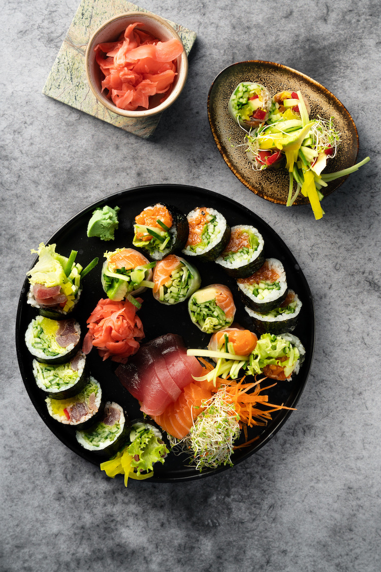 Zestaw sushi i sashimi z dodatkami, ułożony na czarnym talerzu, obok miseczka z marynowanym imbirem i rolki w papierze ryżowym na brązowym talerzu, całość na szarym tle.