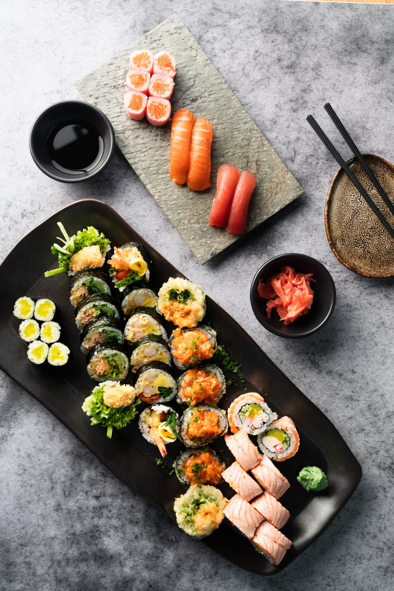 Zestaw sushi: rolki z łososiem, tuńczykiem i warzywami, podane na czarnym talerzu i kamiennej desce, z sosem sojowym, imbirem i pałeczkami na szarym tle.