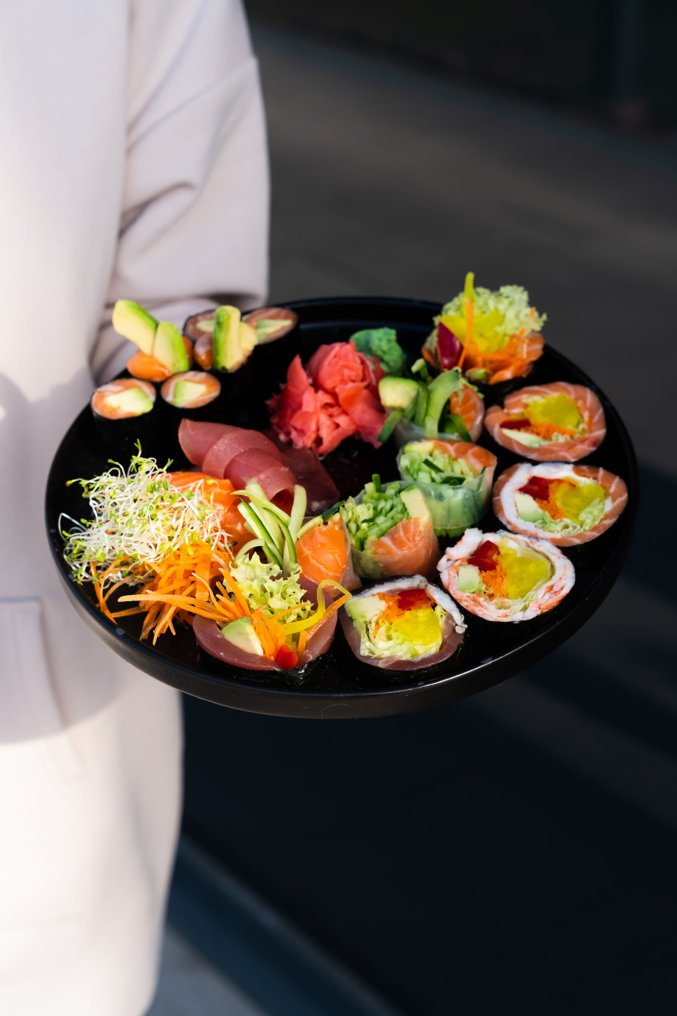 Osoba trzyma czarny talerz z różnorodnym zestawem sushi i sashimi, w tym rolki z łososiem, tuńczykiem, awokado i warzywami, udekorowane imbirem i kiełkami.