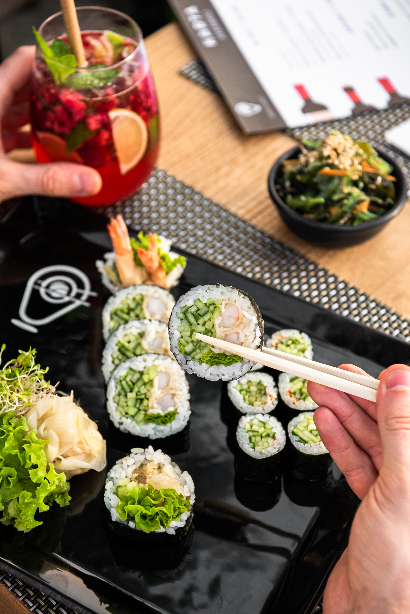 Czarny talerz z sushi maki z krewetką i ogórkiem, pałeczki w dłoni, napój owocowy z miętą i limonką oraz sałatka z wodorostów w tle.