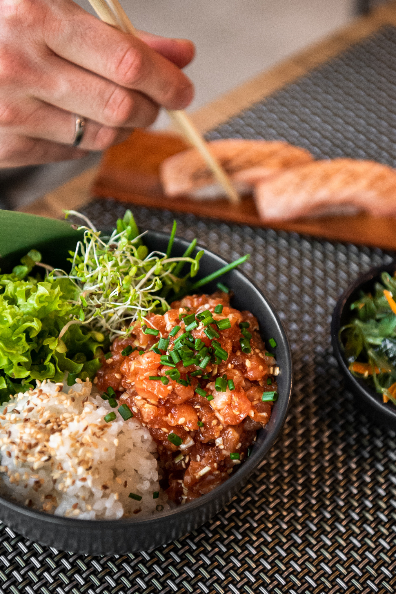 Poke bowl z łososiem, ryżem, sałatą, kiełkami i szczypiorkiem w czarnej misce, trzymana pałeczkami przez osobę z obrączką, na tle deski z łososiem i miski z wodorostami.