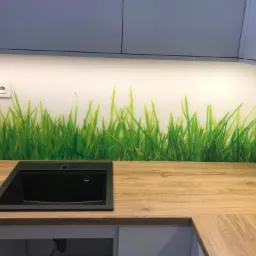 Kuchnia z blatem drewnopodobnym i czarnym zlewem podwieszanym, ściana nad blatem pokryta szkłem z motywem trawy, szare szafki wiszące.