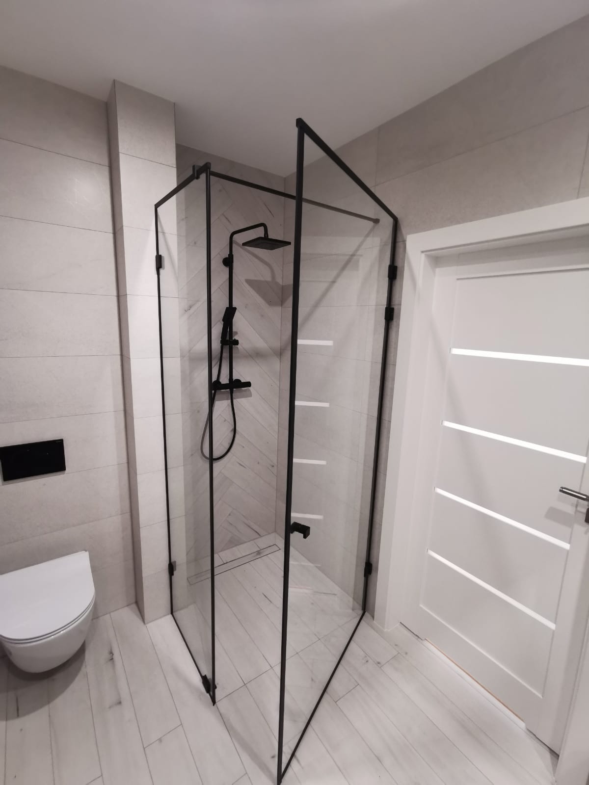 Nowoczesna kabina prysznicowa z czarnymi profilami i deszczownicą w łazience z białymi płytkami imitującymi drewno, widok na drzwi i toaletę podwieszaną.