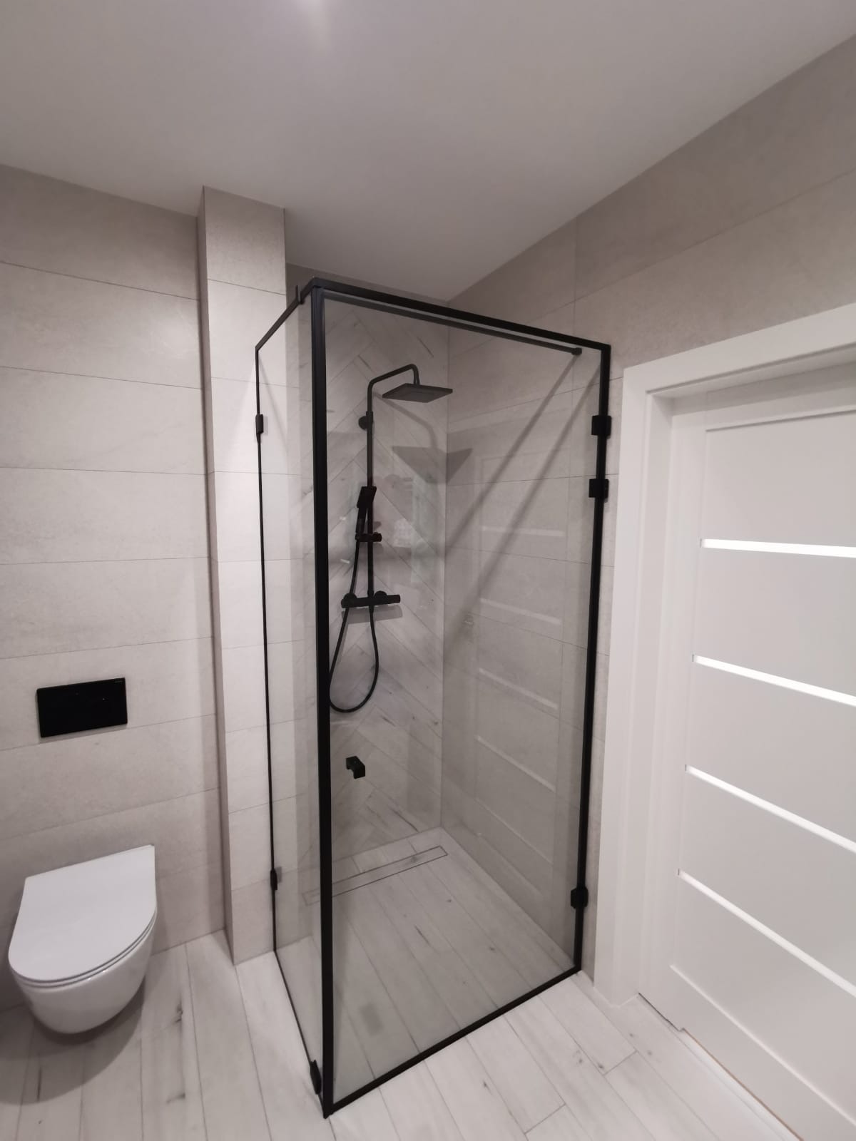 Nowoczesna kabina prysznicowa ze szklanymi ścianami i czarnymi profilami w łazience z jasnymi płytkami imitującymi drewno.