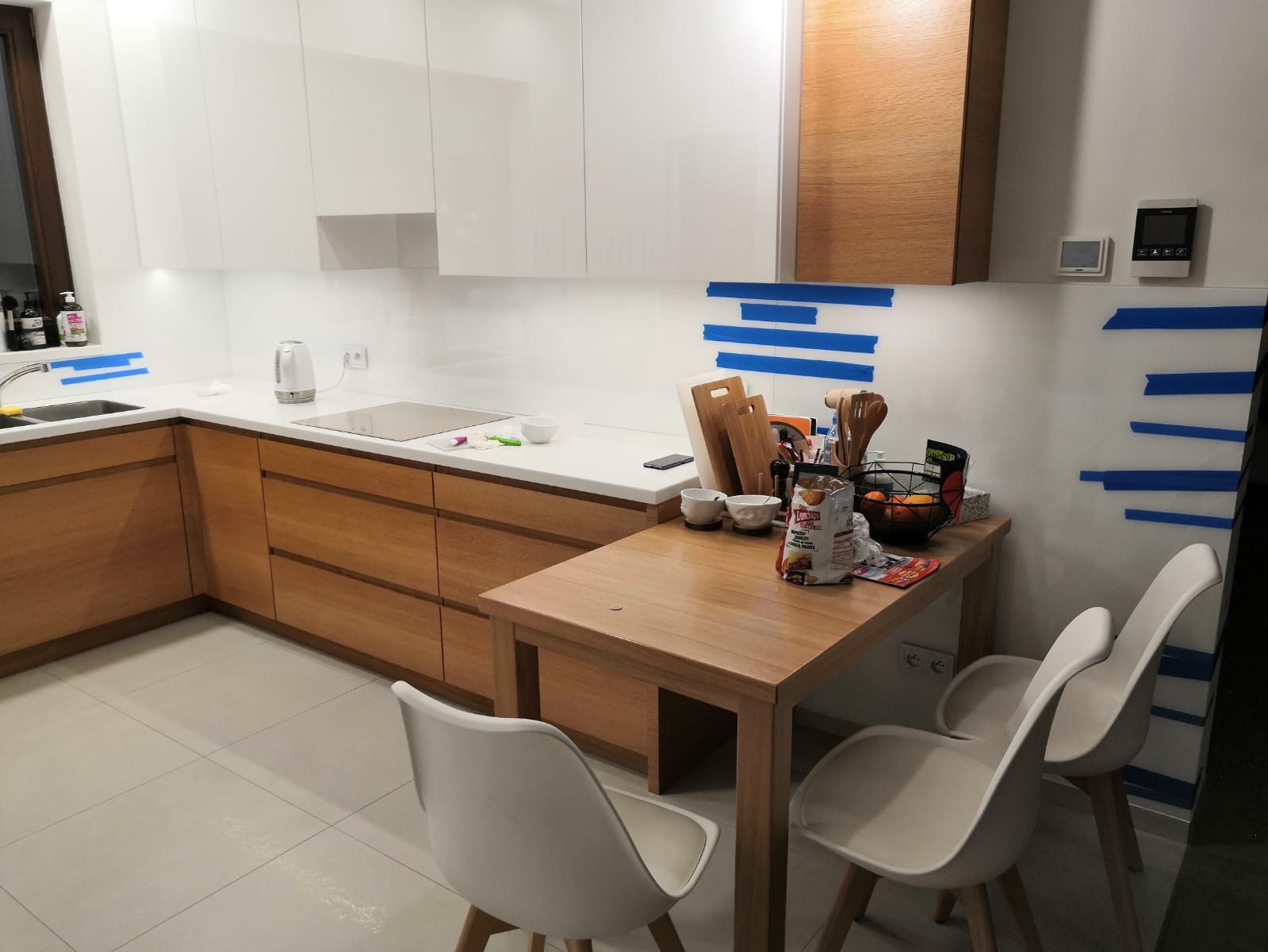 Nowoczesna kuchnia w trakcie wykończenia: białe szafki górne, drewniane szafki dolne, biały blat, zabezpieczona taśmą szklana ściana nad blatem, drewniany stół i białe krzesła.