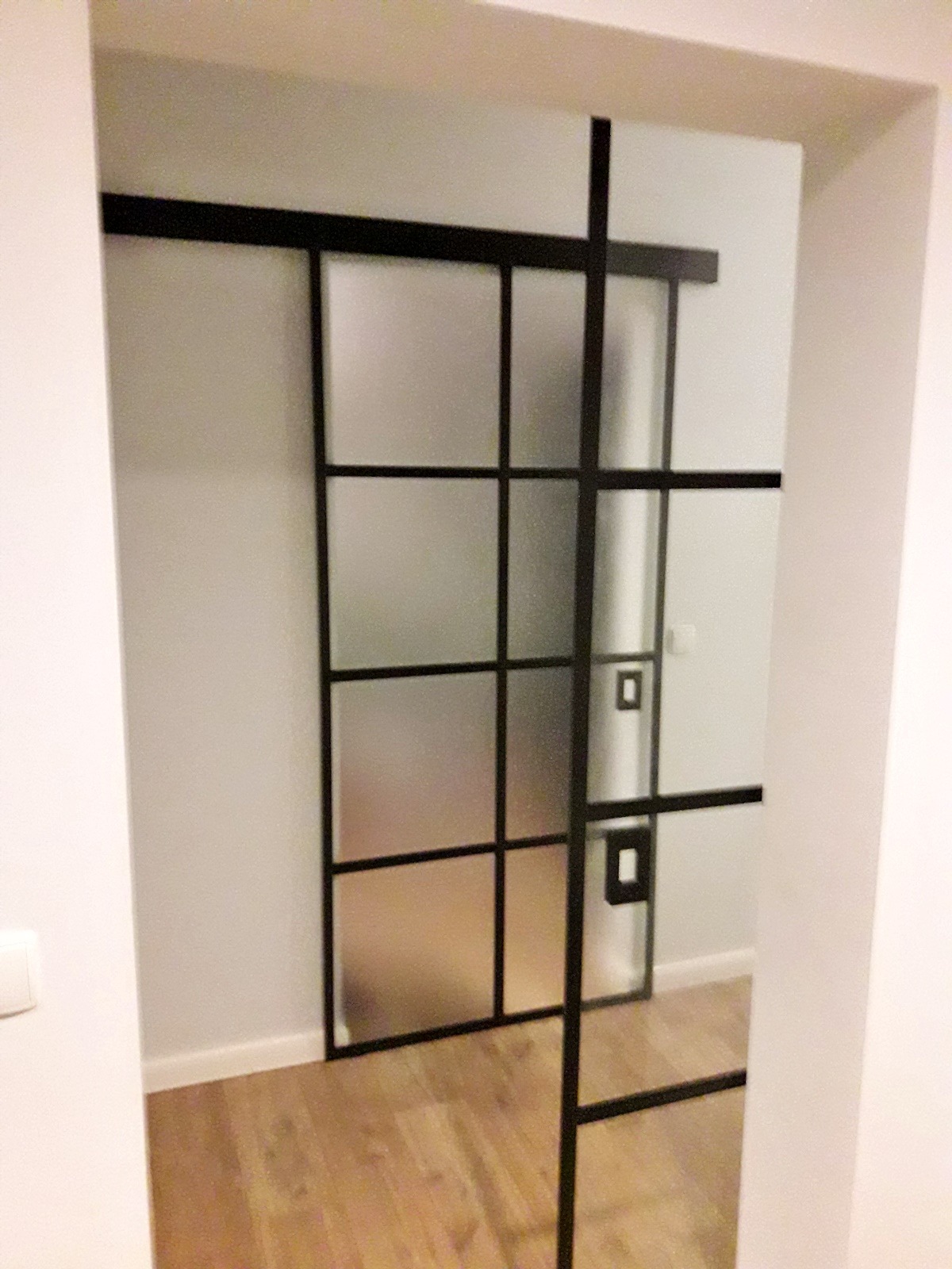 Przesuwne drzwi wewnętrzne z czarnymi szprosami i matowym szkłem, widok z boku na drewnianą podłogę w tle.