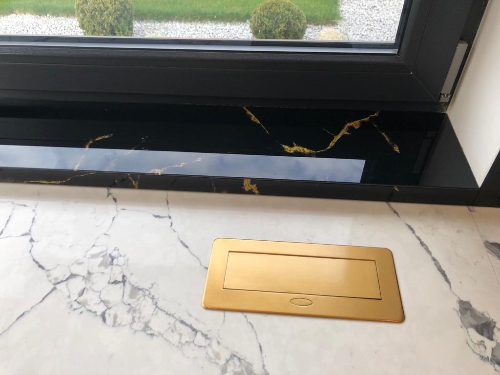 Czarny parapet z żyłkowaniem w kolorze złota, okno z czarną ramą i marmurowy blat z wbudowanym, złotym gniazdkiem elektrycznym.