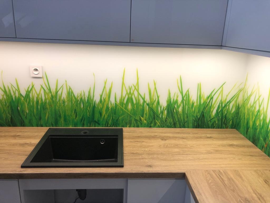 Kuchnia z blatem drewnopodobnym i czarnym zlewem podwieszanym, ściana nad blatem pokryta szkłem z motywem trawy, szare szafki wiszące.